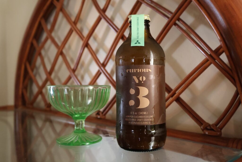 Curious Elixirs No. 3 (Juniper Cucumber Collins) next to a green glass on a glass table with a lattice wood background