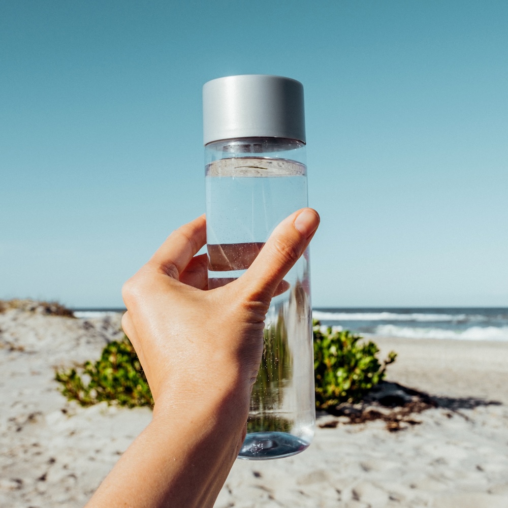 Image of a female hand holding a glass water bottle with a metal top at the beach. Meant to convey ways to reduce plastic like using a reuseable water bottle